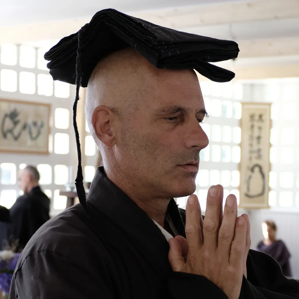 Un moine zen chante un sutra en posture de gassho avec son kesa sur la tête.